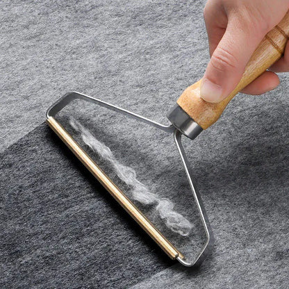 A close-up of a hand holding a lint roller with a light wood handle and metal frame, rolling over gray fabric to show the removal of white lint.